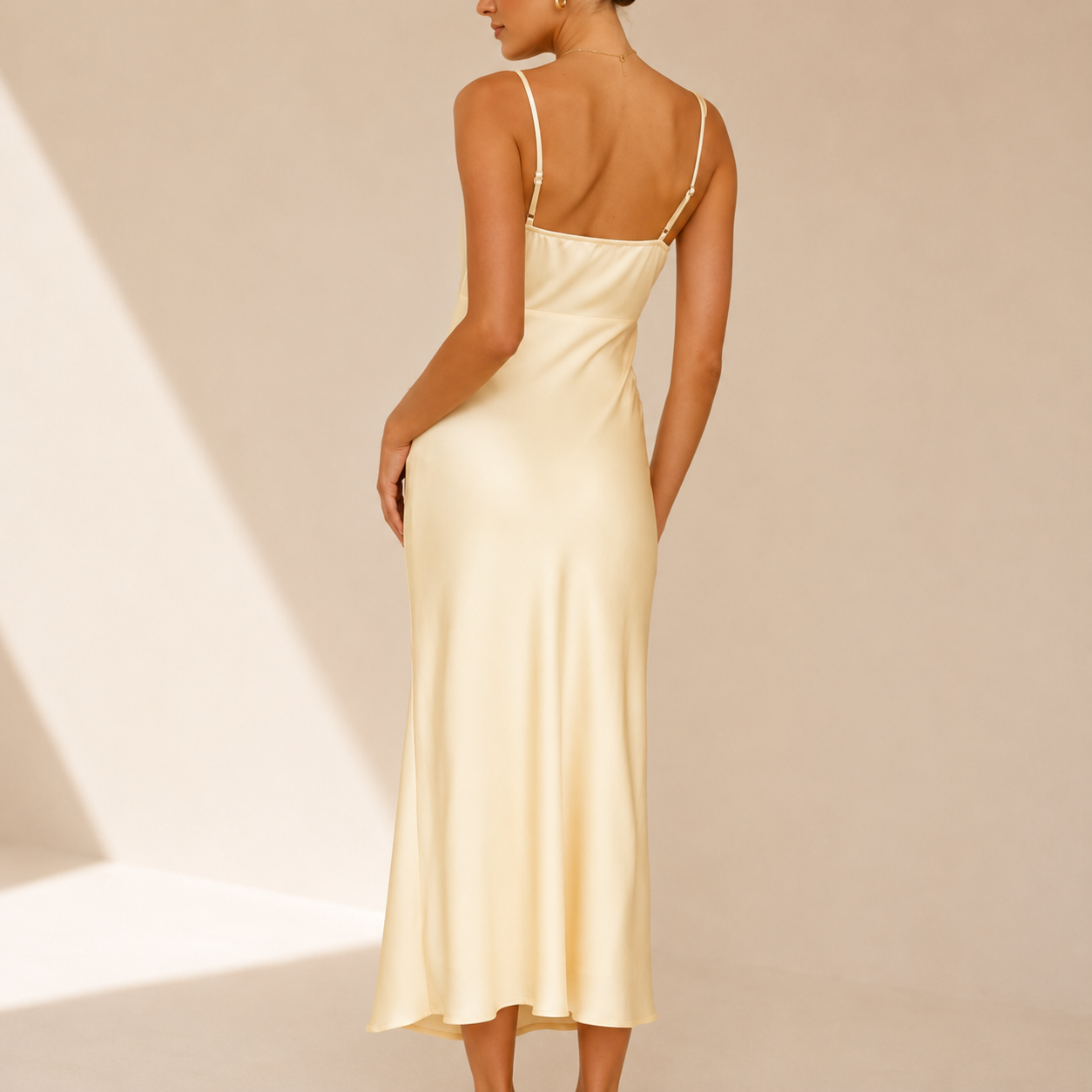 A woman wearing a light cream satin midi slip dress, shown from the back against a warm beige background with soft diagonal shadows.
